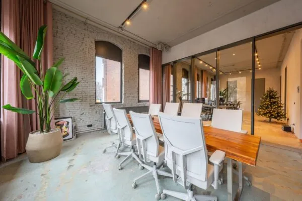 Vergaderruimte aan de Admiraal de Ruijterweg met een houten tafel, witte bureaustoelen, grote kamerplant en zicht op een verlichte kerstboom in de aangrenzende ruimte.